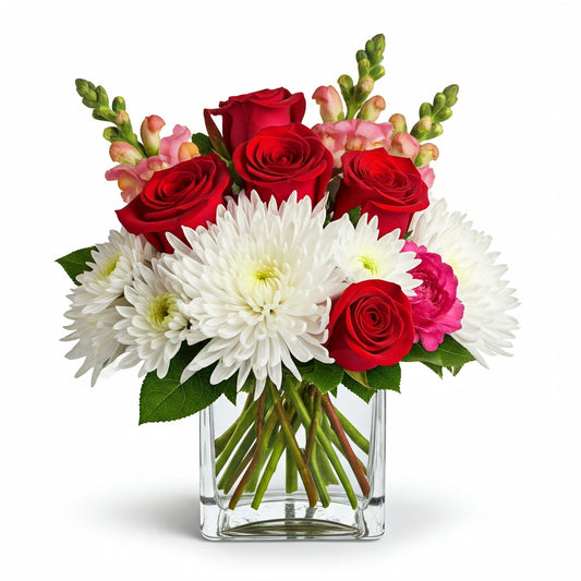 A lush Romantic Bliss flower arrangement in a square glass vase, featuring red roses, white chrysanthemums, and pink snapdragons, accented with subtle greenery. Perfect for Valentine's Day.